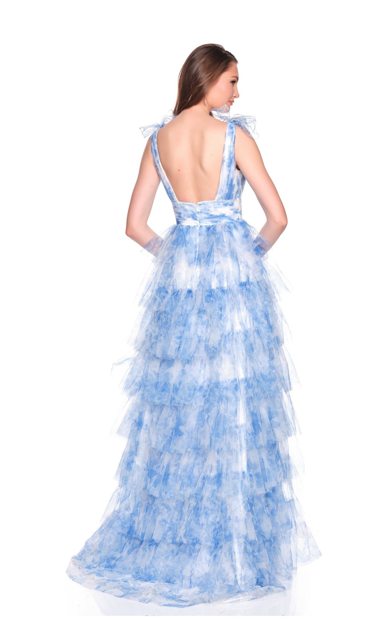 A woman stands facing away from the camera in a sleeveless, floor-length blue Dave and Johnny 12793 prom dress with white ruffles and a deep V-back, set against a white background.